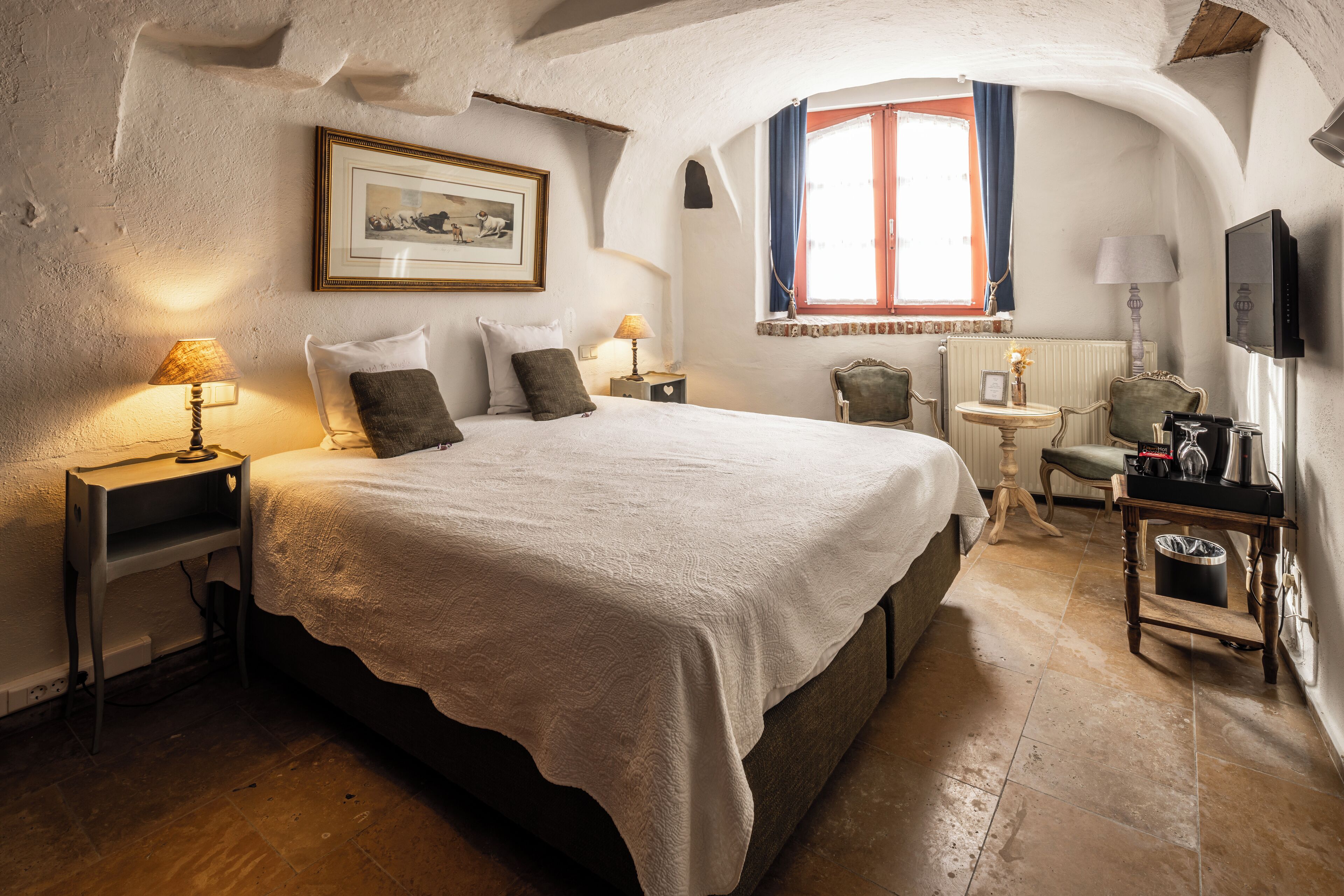 Ein Schlafzimmer mit einem großen Bett, zwei Nachttischen, einem Sessel, einem kleinen Tisch, einem Fernseher und einem Fenster mit Vorhängen.