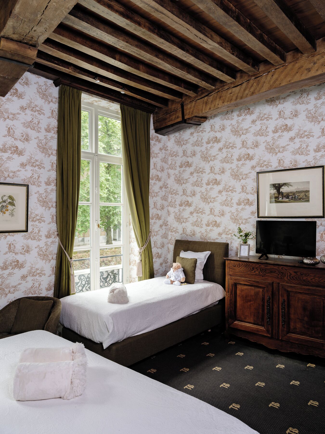 Un dormitorio con techo de madera, una cama con edredón blanco, un televisor sobre un mueble de madera y una ventana con cortinas verdes.
