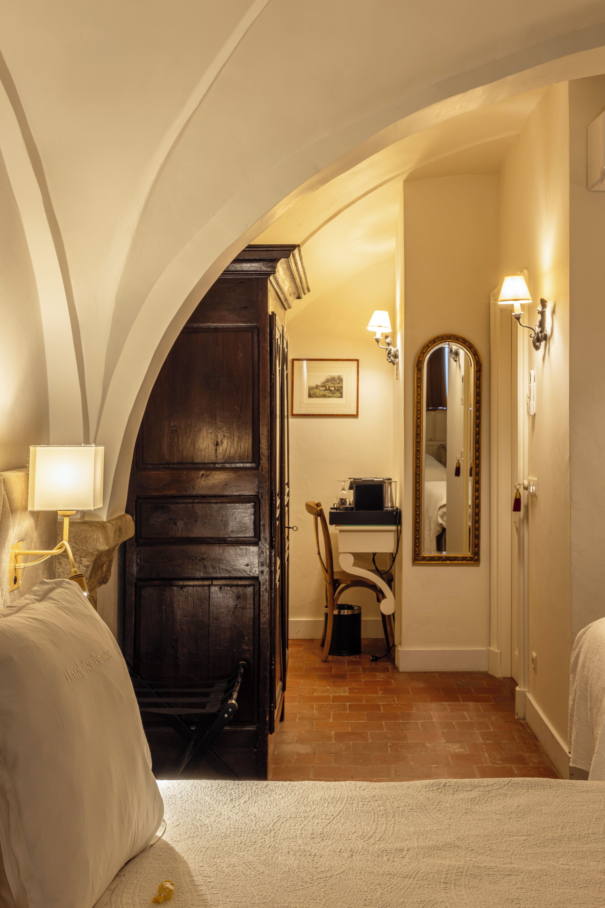 A hotel room with a bed, a desk, a chair, a mirror, and a lamp.