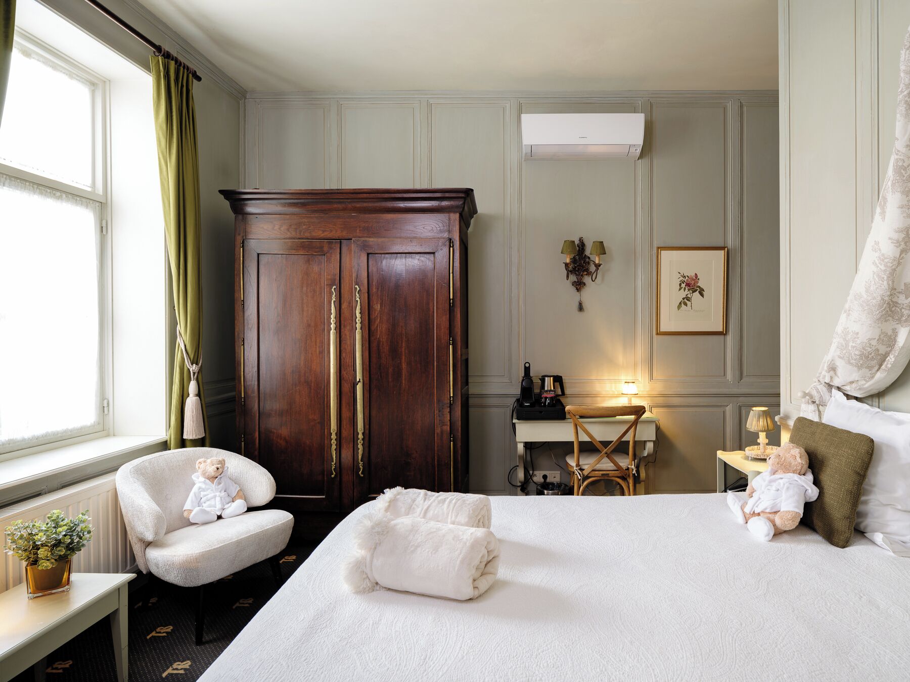 A hotel room with a large bed, two chairs, a desk with a lamp, a wardrobe, and a window with curtains.