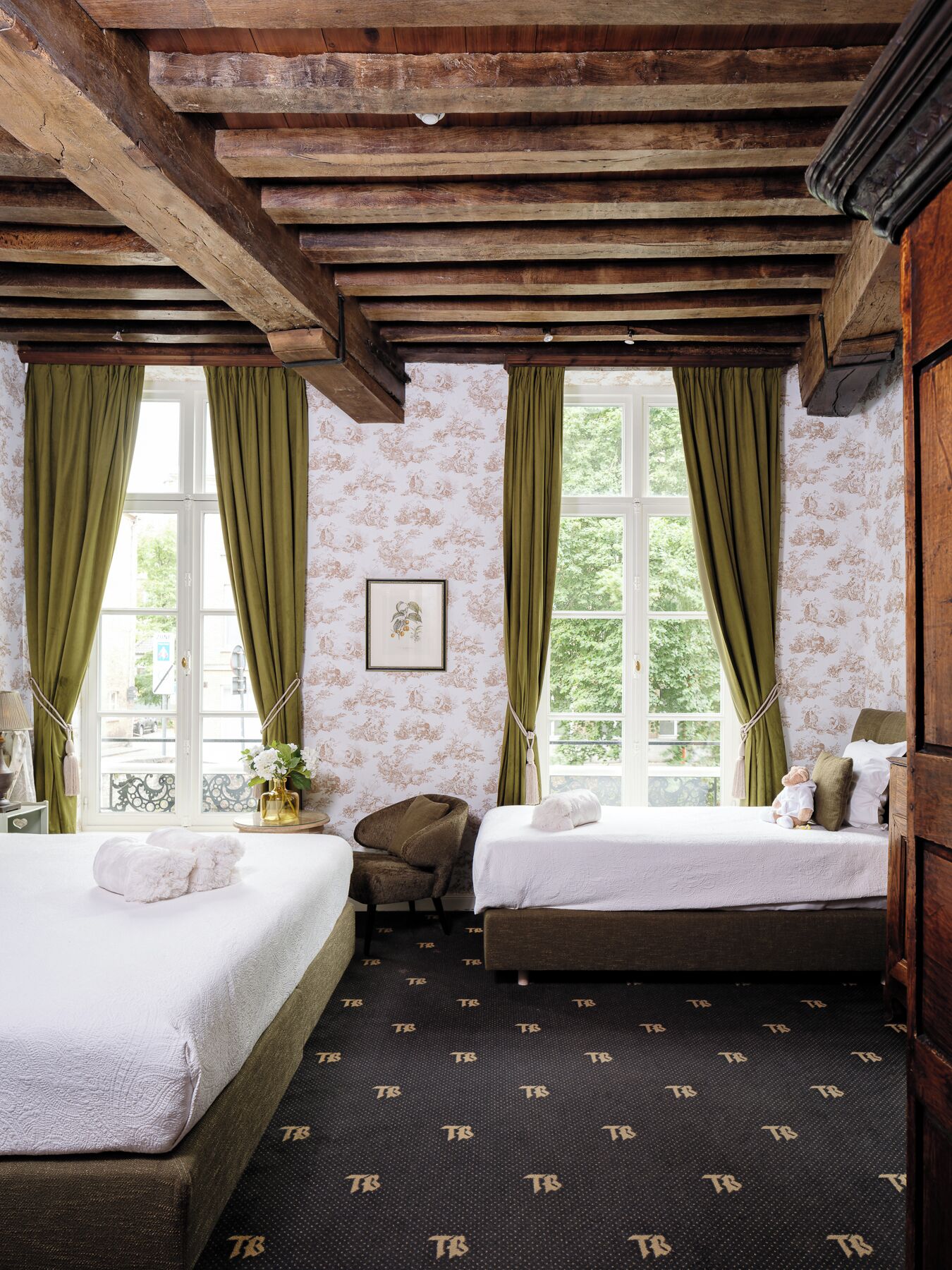 Ein Zimmer mit zwei Betten, einem Sessel, einem Fenster mit Vorhängen und einer Holzbalkendecke.