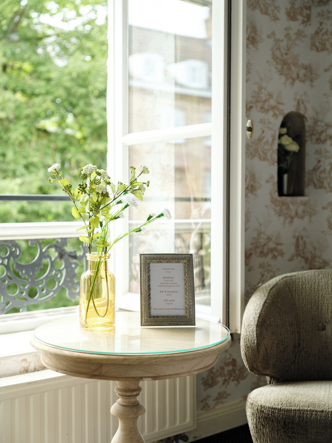 Ein Zimmer mit Fenster, eine Vase mit Blumen, ein gerahmtes Schild mit Frühstückszeiten und ein Stuhl.