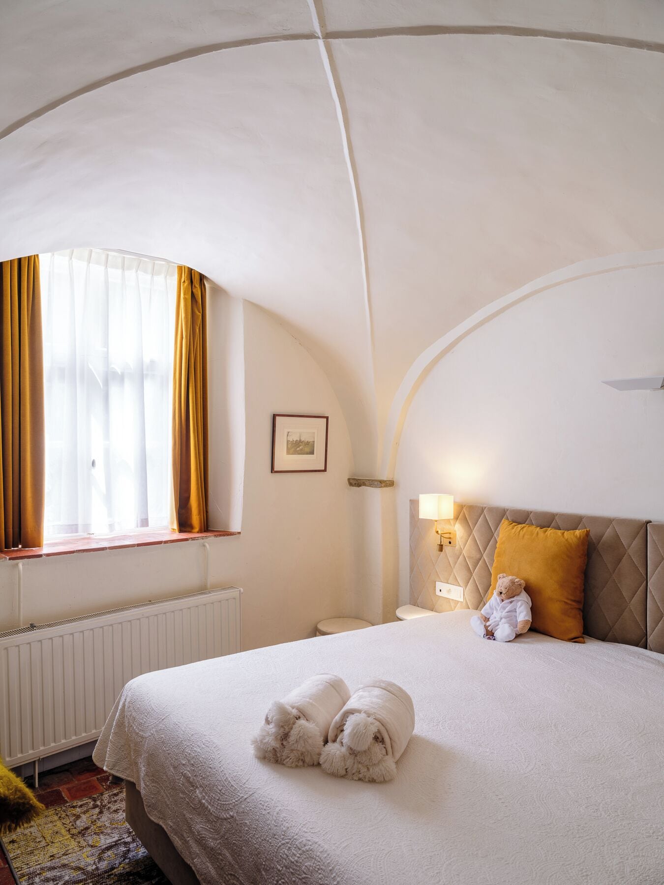 A hotel room with a large bed, a window with curtains, a bedside lamp, and a teddy bear on the bed.