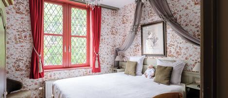 A bedroom with a large bed, a chandelier, a window with red curtains, floral wallpaper, and a bedside table with a lamp.