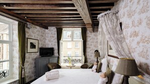Un dormitorio con una cama grande, un televisor, una mesita con un florero y una ventana con cortinas.