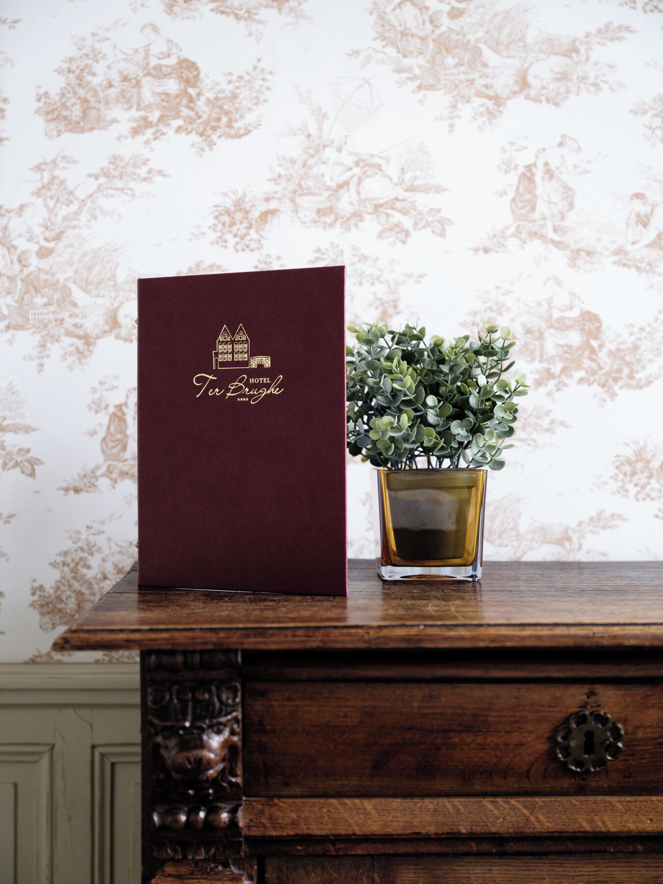 Une carte d’hôtel de couleur bordeaux avec des lettres dorées, et une petite plante en pot posée sur une surface en bois.