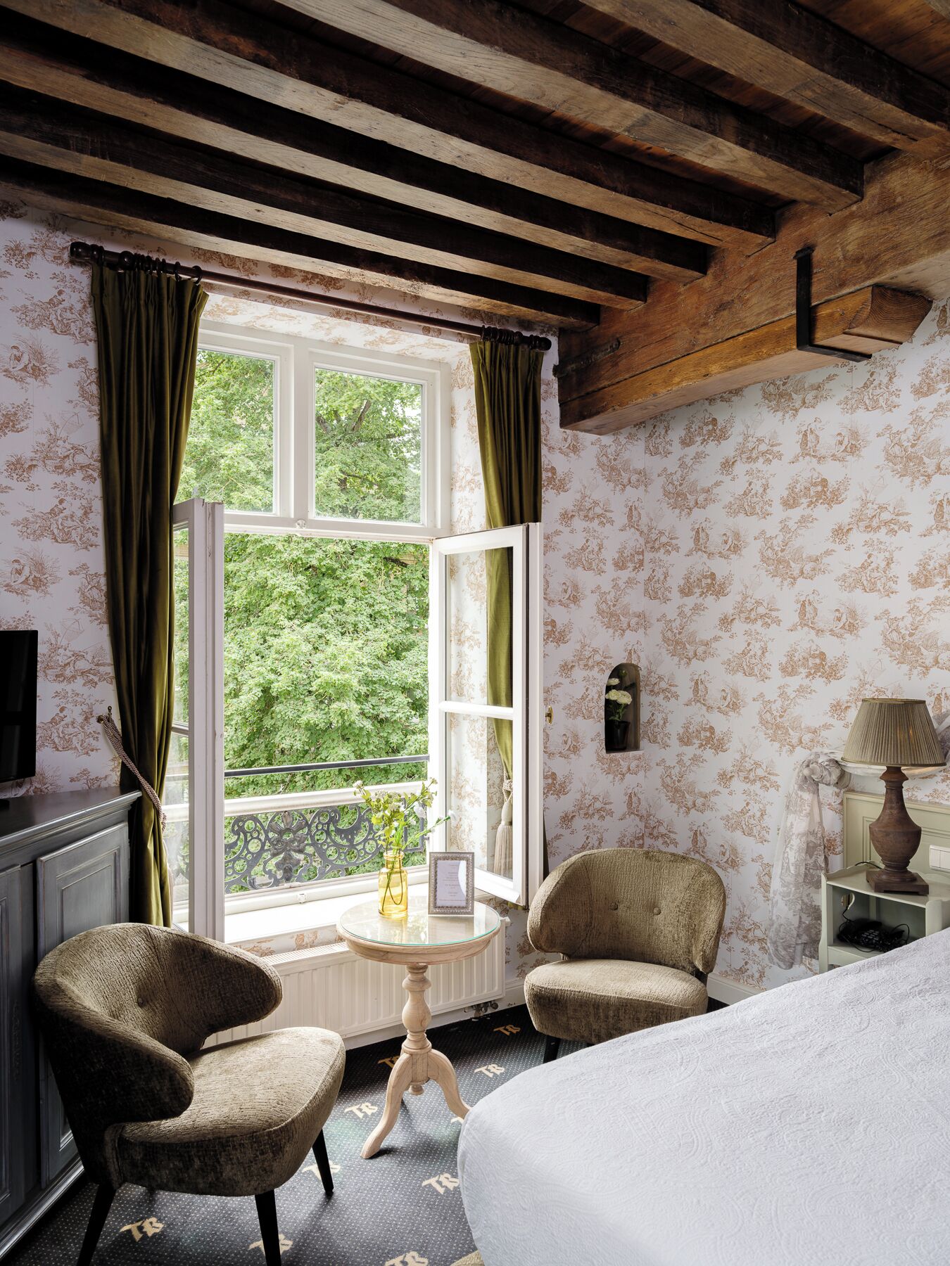 Une pièce comprenant un lit, deux fauteuils, une petite table, une lampe et une fenêtre avec des rideaux.
