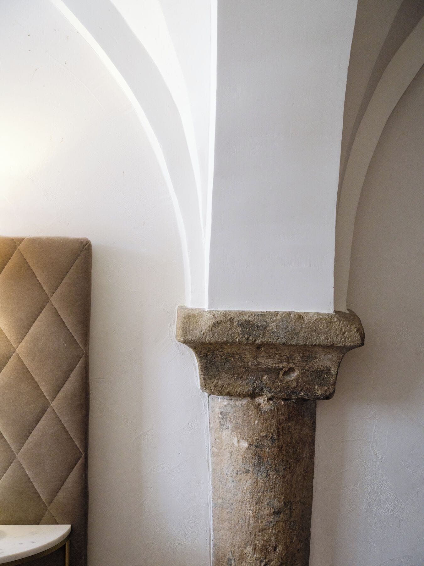 Um pilar de pedra com topo arredondado, uma cadeira estofada bege e uma parede branca com um desenho em arco.