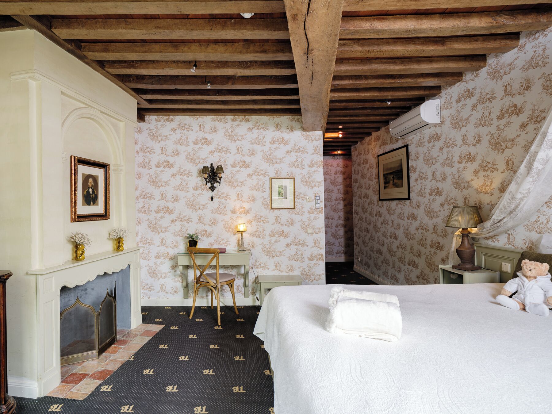 Un dormitorio con cama, una silla, escritorio, chimenea y una pared con cuadros enmarcados.
