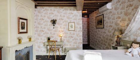 A bedroom with a bed, a chair, a desk, a fireplace, and a wall with framed pictures.