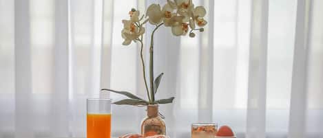 Une table dressée pour le petit-déjeuner, avec des croissants, des fruits, un verre de jus d’orange et un dessert, placée près d’une fenêtre ornée de rideaux légers.