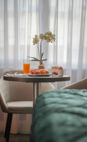 Desayuno con croissants, fruta, un vaso de zumo de naranja y un postre sobre una mesa junto a una ventana con cortinas traslúcidas.