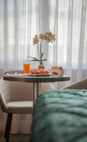 A breakfast setup with croissants, a fruit, a glass of orange juice, and a dessert on a table by a window with sheer curtains.
