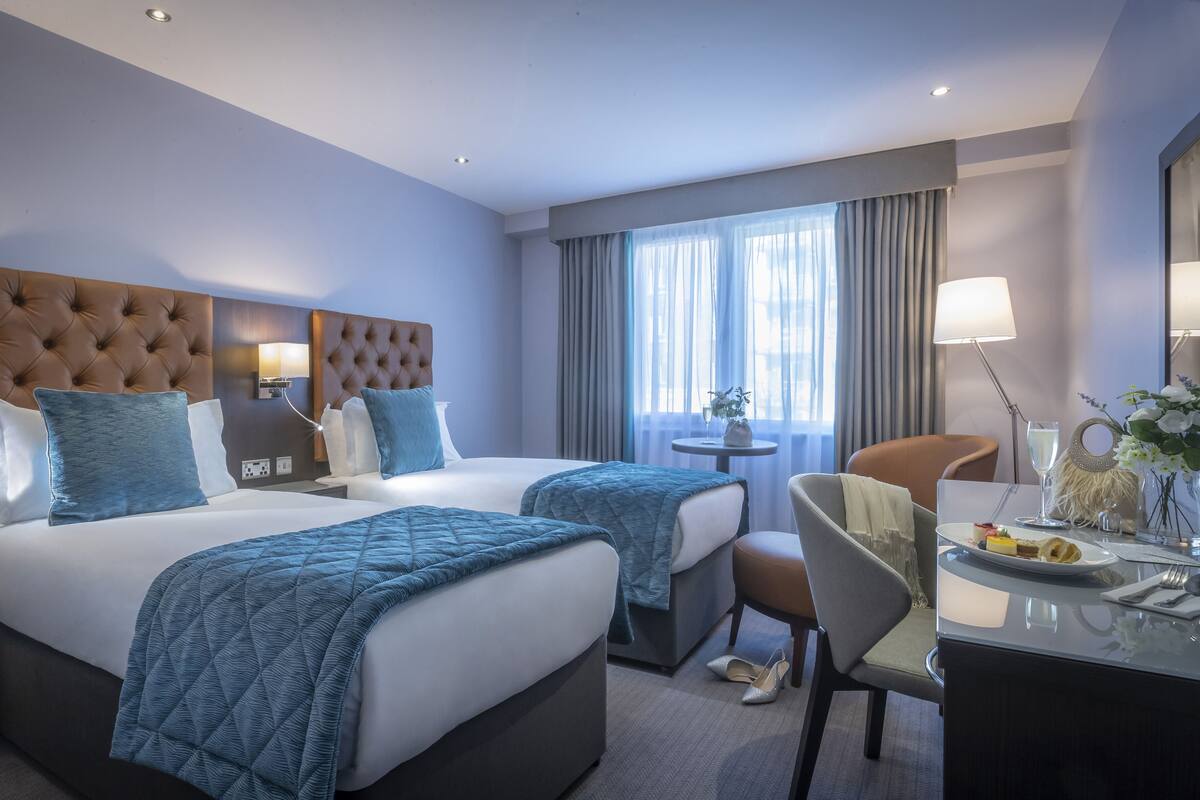 A hotel room with two beds, a desk, a chair, a lamp, and a view of the city through large windows.