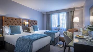 A hotel room with two beds, a desk, a chair, a lamp, and a view of the city through large windows.