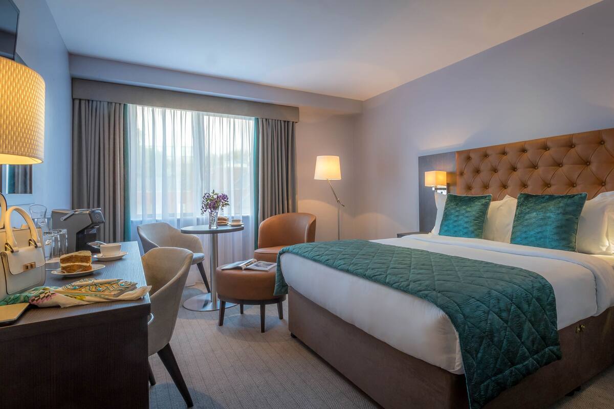 A hotel room with a large bed, a desk with a chair, a small table with a vase of flowers, and a lamp.