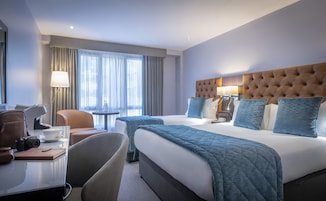 A hotel room with a large bed, a desk with a chair, a lamp, and a window with curtains.
