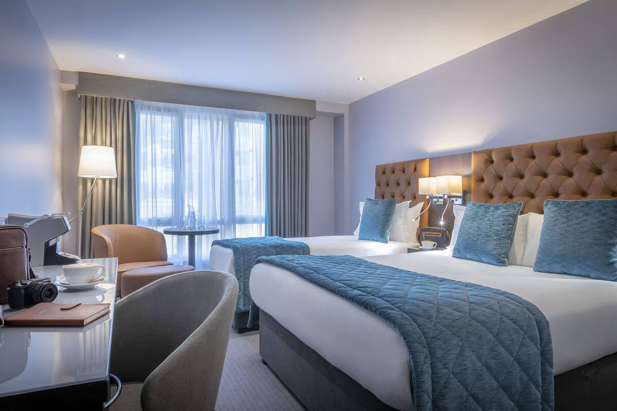 A hotel room with a large bed, two bedside tables, a desk with a chair, a lamp, and a window with curtains.