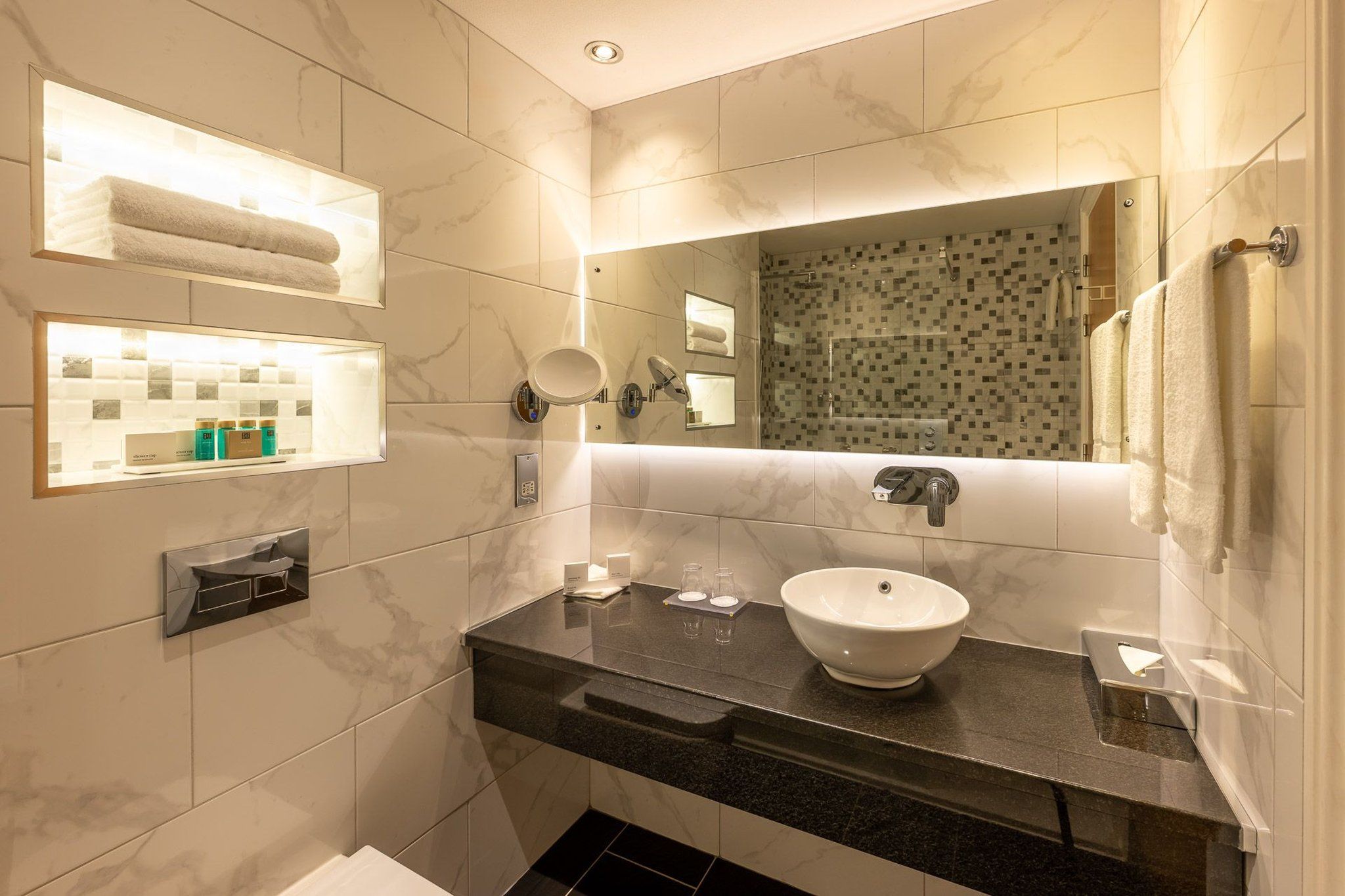 Un baño moderno con pared de azulejos de mármol, encimera negra, lavabo blanco y zona de ducha con mampara de vidrio.