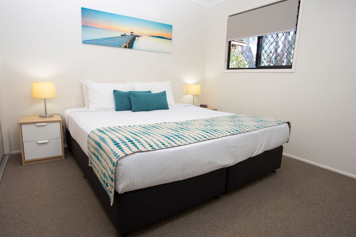 Une chambre à coucher bien aménagée, avec un grand lit, deux tables de chevet avec des lampes, une fenêtre donnant sur un beau paysage et un tableau accroché au mur.