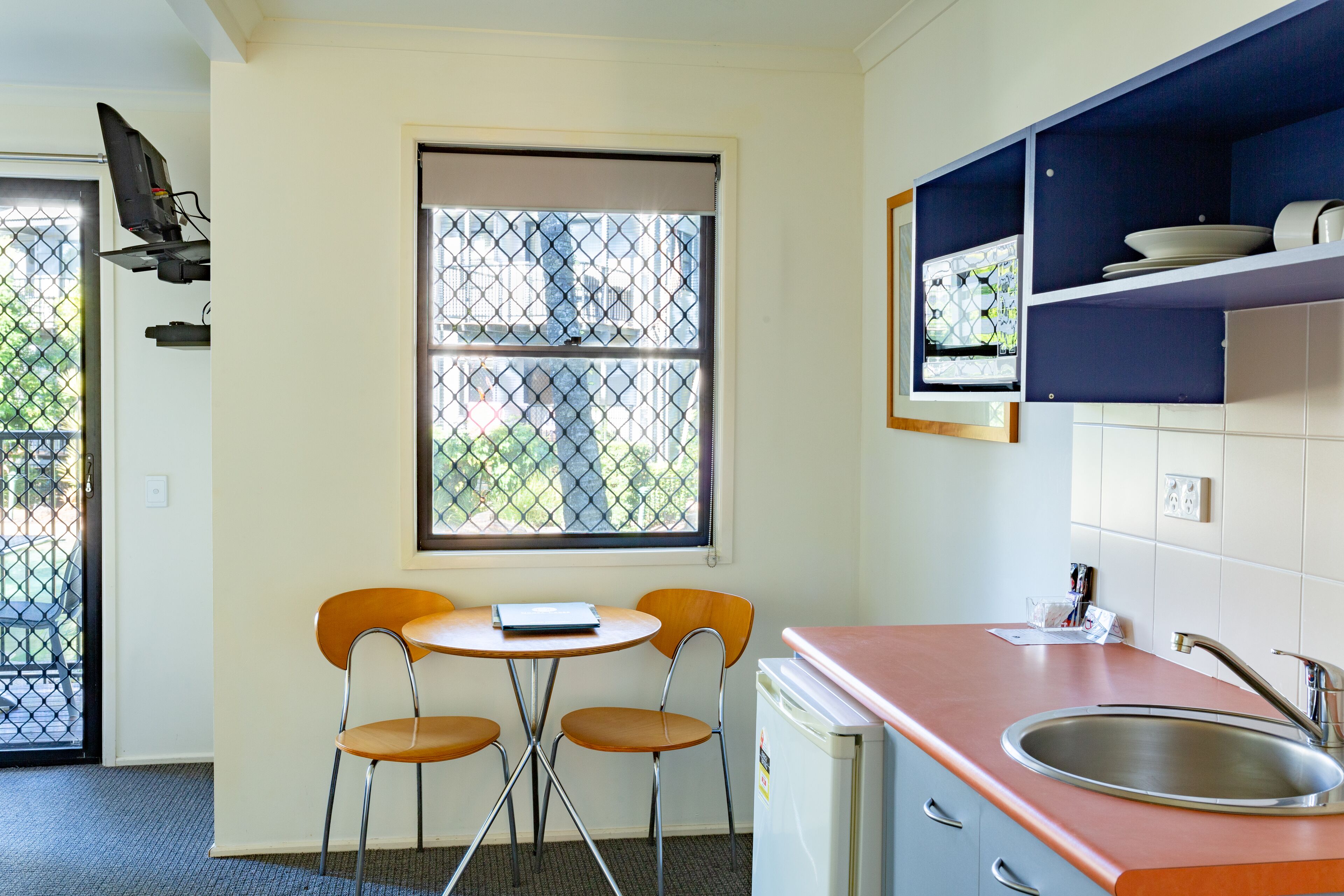 Cocina compacta con lavamanos, un refrigerador pequeño, un ventanal con vista a edificios y una mesa con dos sillas.