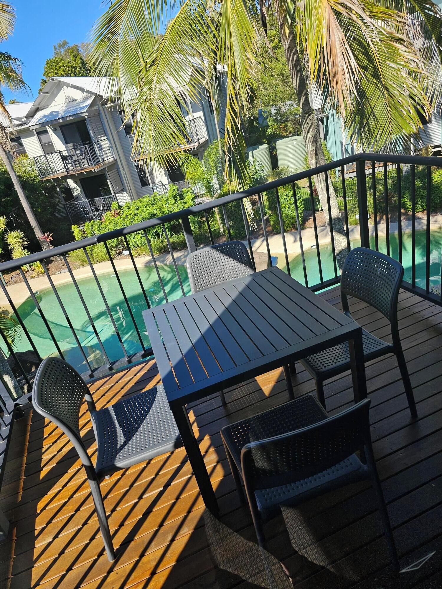 A wooden deck with a table and chairs, overlooking a swimming pool and lush greenery.