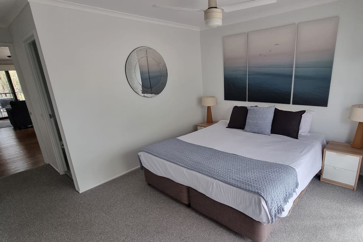 Une chambre moderne avec un grand lit, un ventilateur de plafond, un miroir fixé au mur et une vue sur le ciel par une fenêtre.