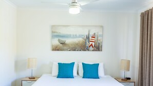 A hotel room with a bed, two nightstands, a ceiling fan, a painting of a beach scene, and a window with curtains.
