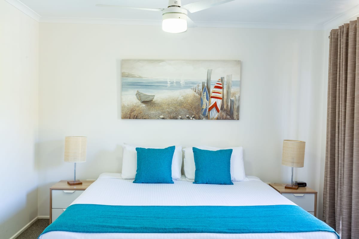 Une chambre d’hôtel avec un lit, deux tables de chevet, un ventilateur de plafond, un tableau représentant une scène de plage et une fenêtre avec des rideaux.