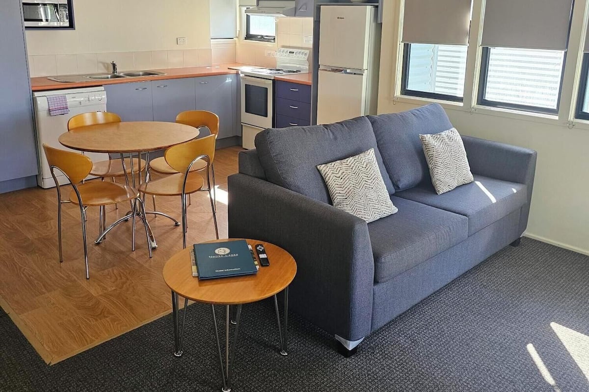 Un espace de vie compact comprenant un coin cuisine, un canapé gris avec des coussins, une petite table à manger avec des chaises et un sol recouvert d’une moquette.