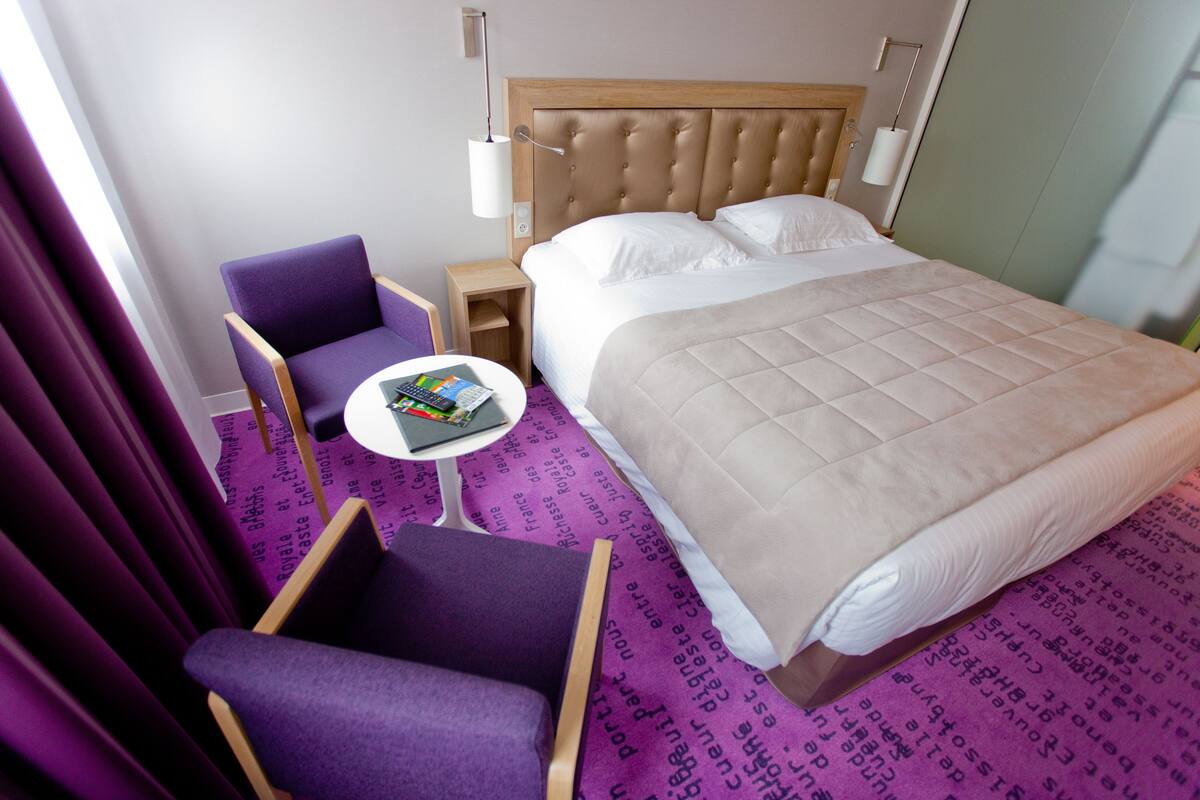 A hotel room with a bed, a purple armchair, a small table with a book and a calculator, and a nightstand with a lamp.