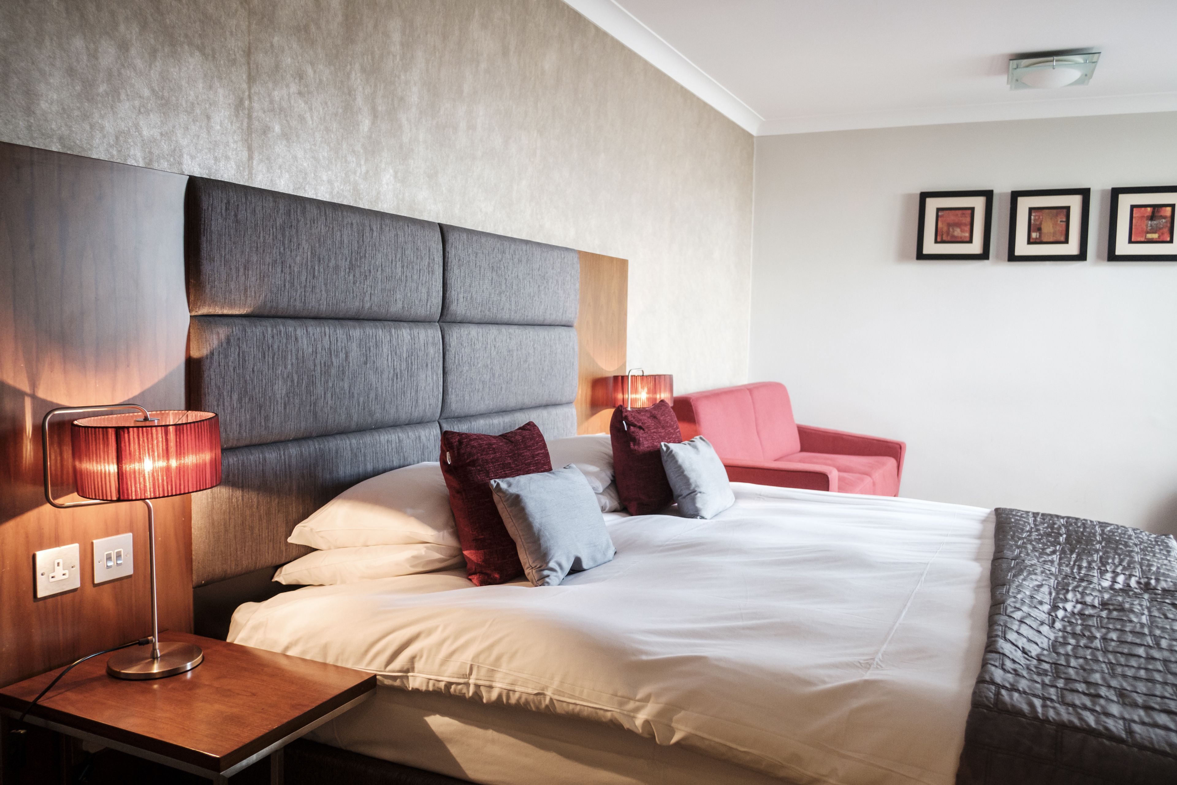 A modern hotel room with a large bed, a wooden nightstand, a red chair, and a wall-mounted lamp.