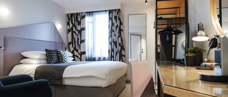 A modern hotel room with a bed, a desk, a chair, a mirror, and a potted plant.