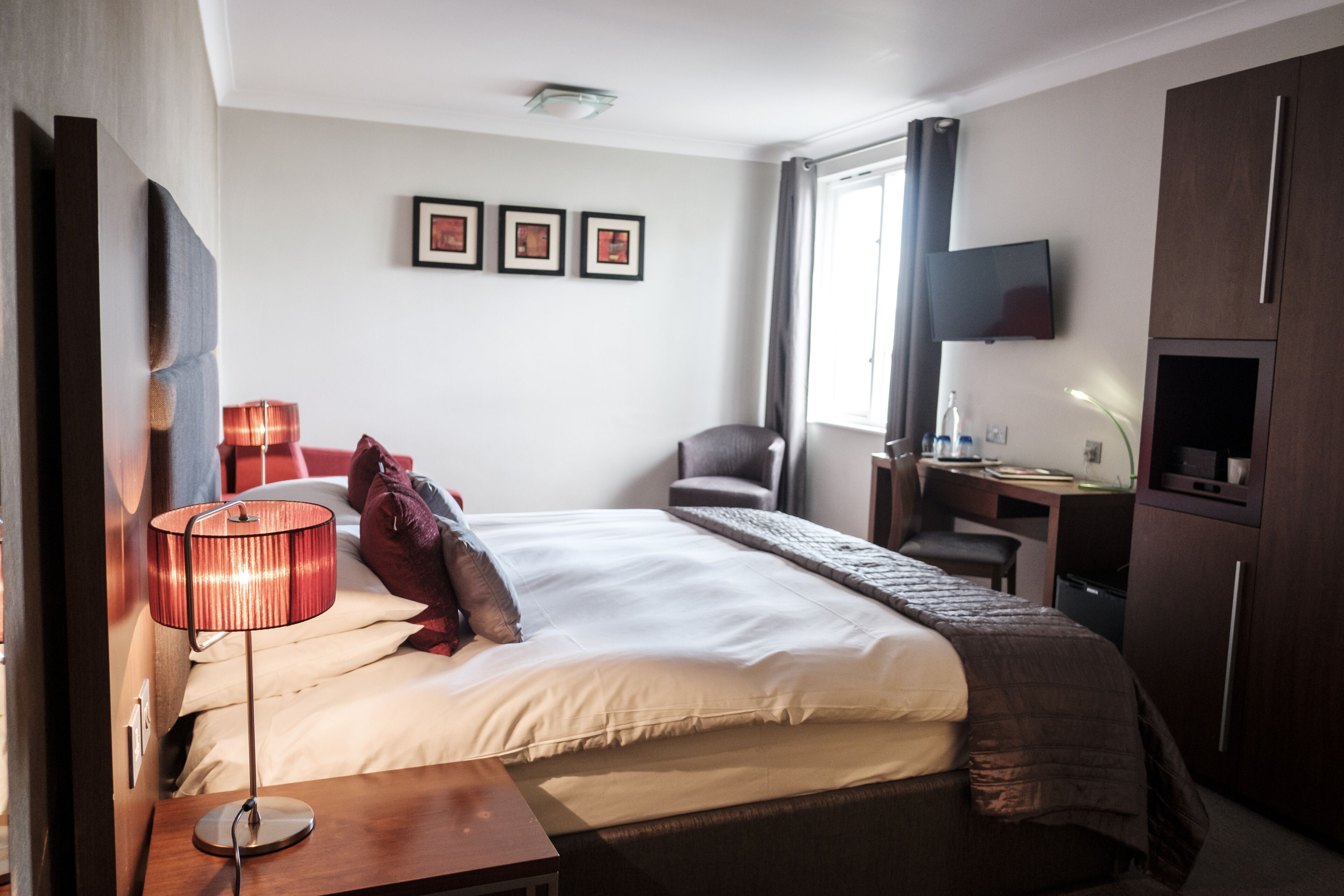 A hotel room with a bed, bedside table, lamp, chair, desk, TV, and wardrobe.