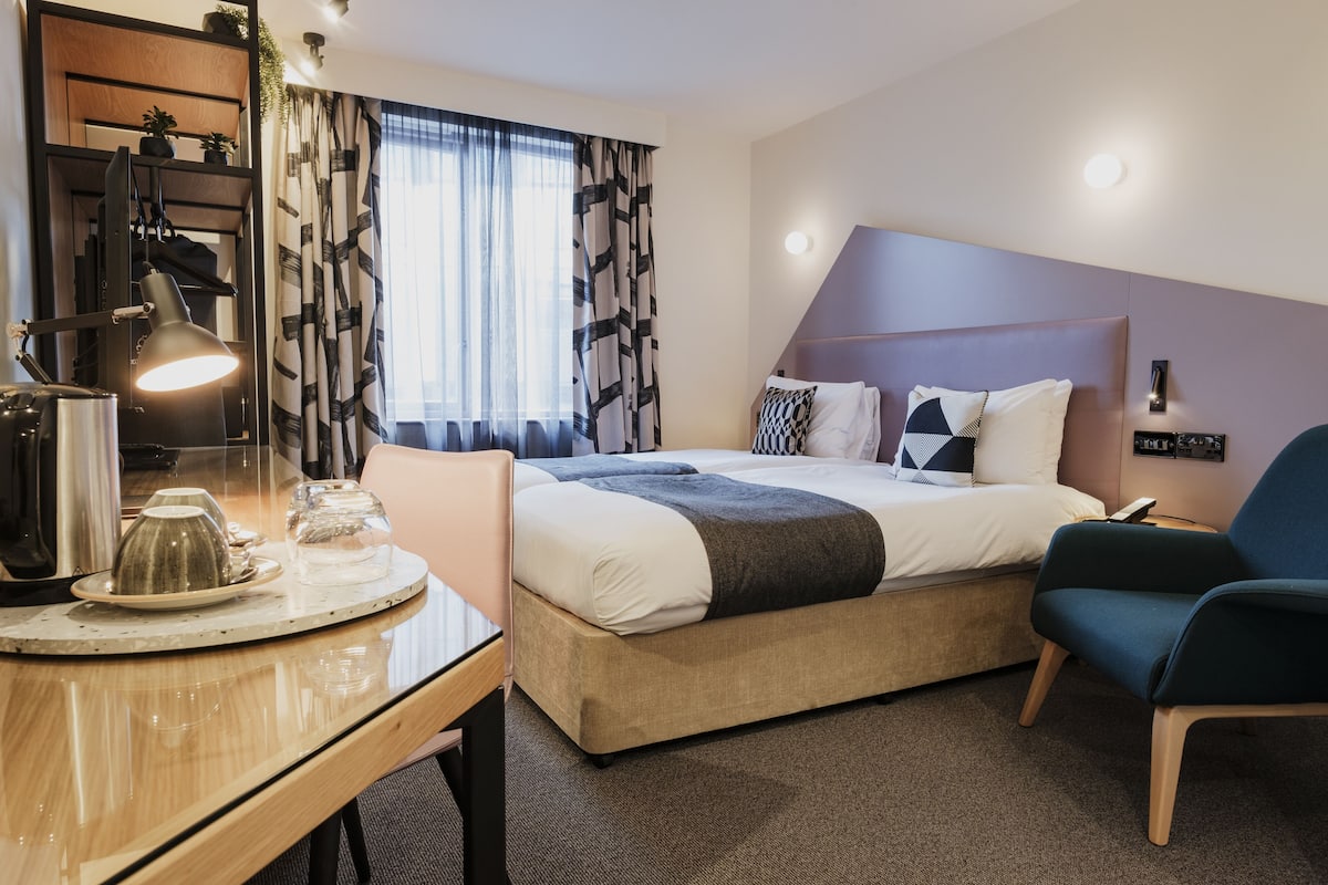 A hotel room with a bed, a desk with a lamp, a chair, and a window with curtains.