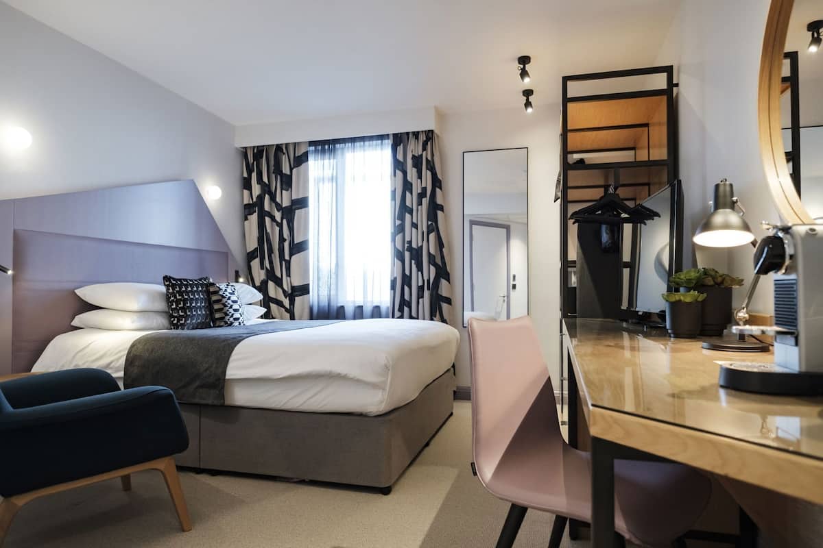 A modern hotel room with a bed, a desk, a chair, a mirror, a lamp, and a coat rack.