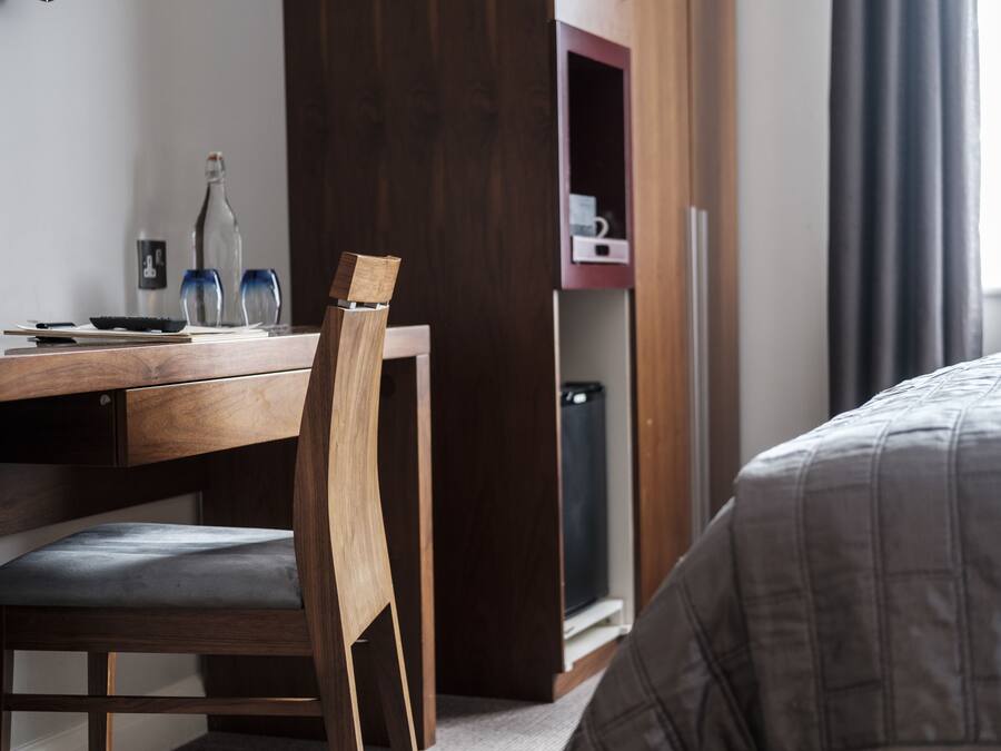 Un escritorio de madera con una silla, una botella y vasos. Se observa una cama con una manta gris.