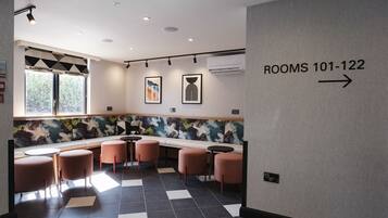 A modern hotel lobby with a checkered floor, a seating area with round tables and chairs, and a sign indicating rooms 101-122.