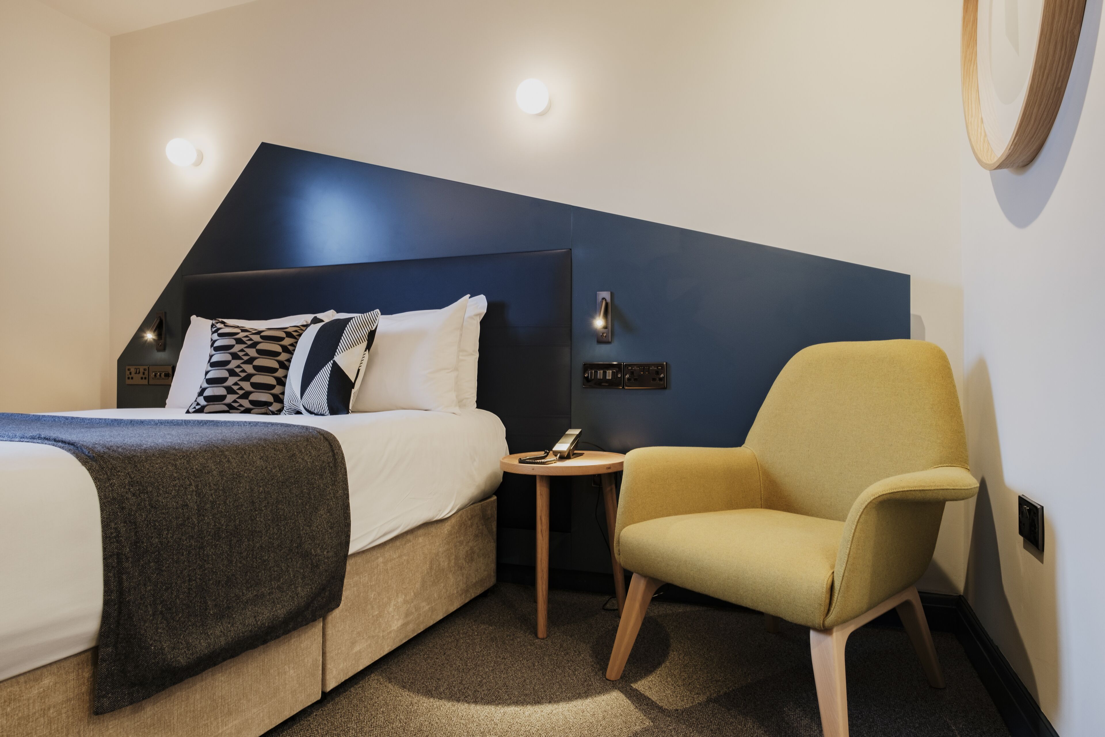 A modern hotel room with a bed, a small round table, a chair, and a unique wall design.