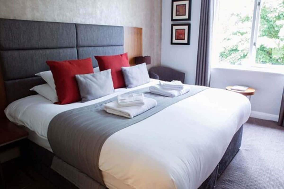 A neatly made bed with a grey headboard, red and white pillows, and folded white towels. There is a small bedside table with a book and a lamp. The room has a large window with a view of greenery outside.