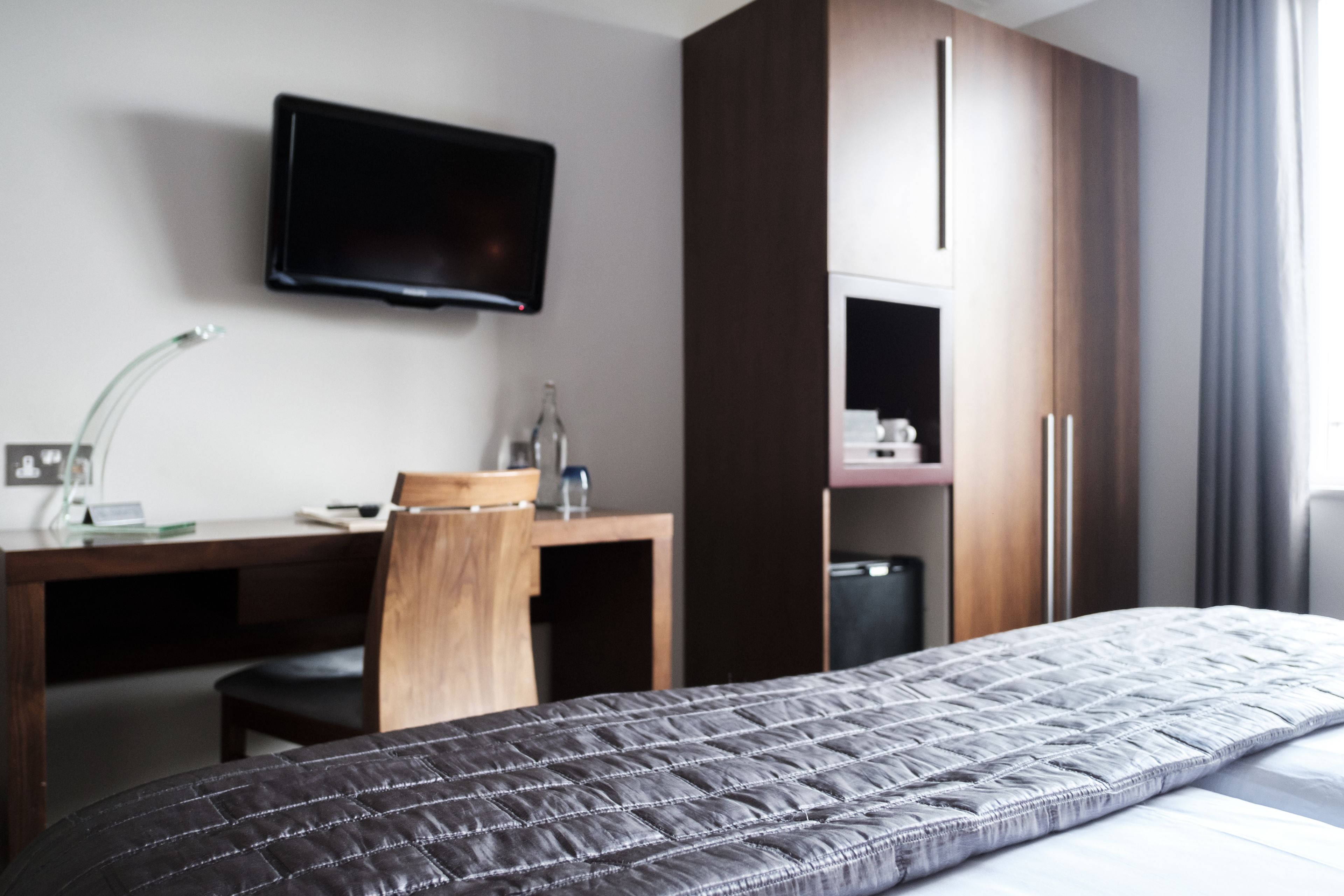 A hotel room with a bed, a desk, a chair, a television mounted on the wall, and a wardrobe.
