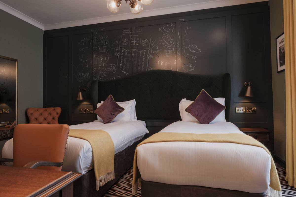 A hotel room with two beds, a large dark headboard, a chair, bedside tables, and a patterned floor.