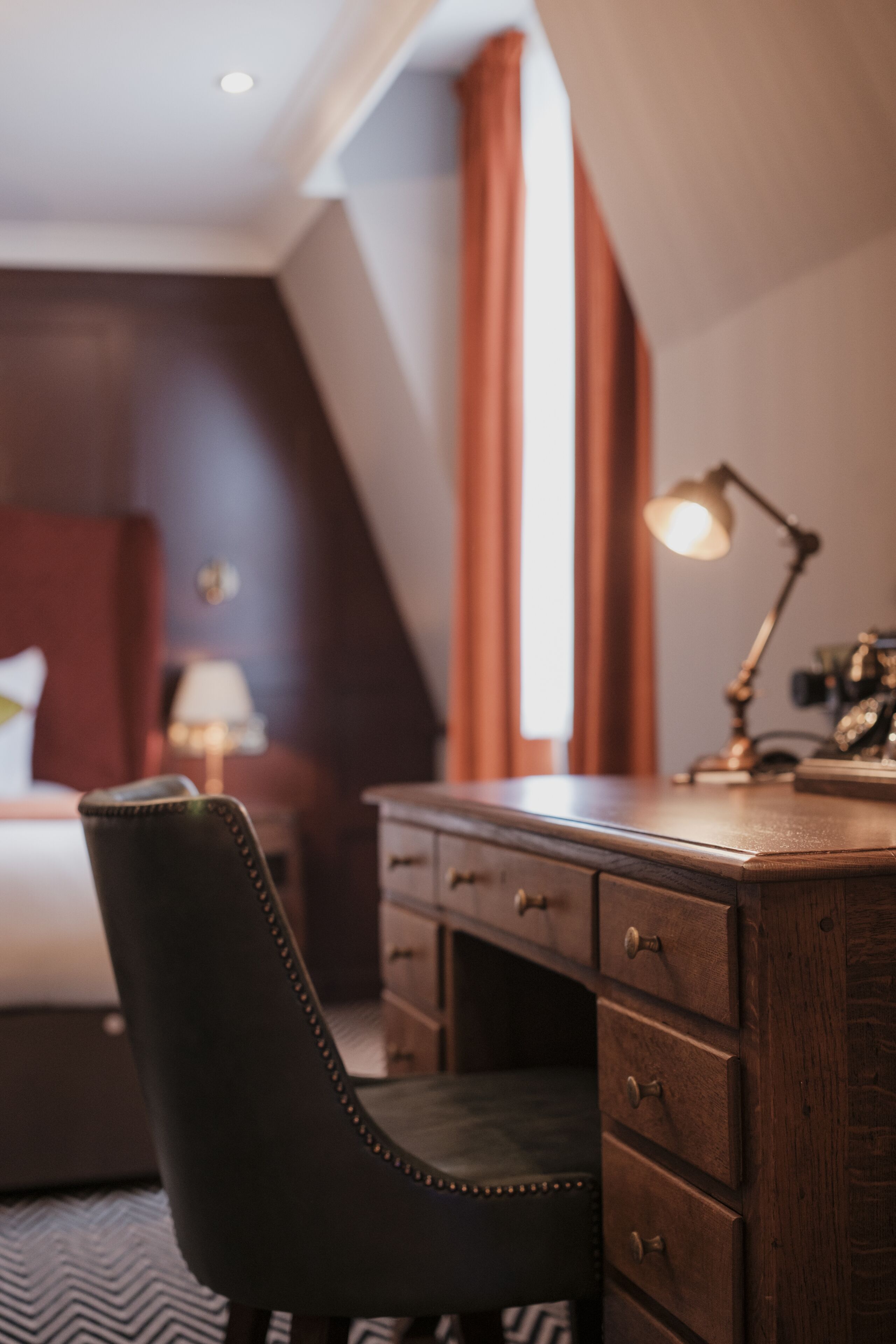 Hotellihuone, jossa on sänky, kirjoituspöytä laatikoineen, lamppu ja tuoli.