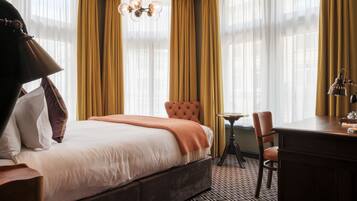 A hotel room with a bed, a desk, a chair, a lamp, and a chandelier.