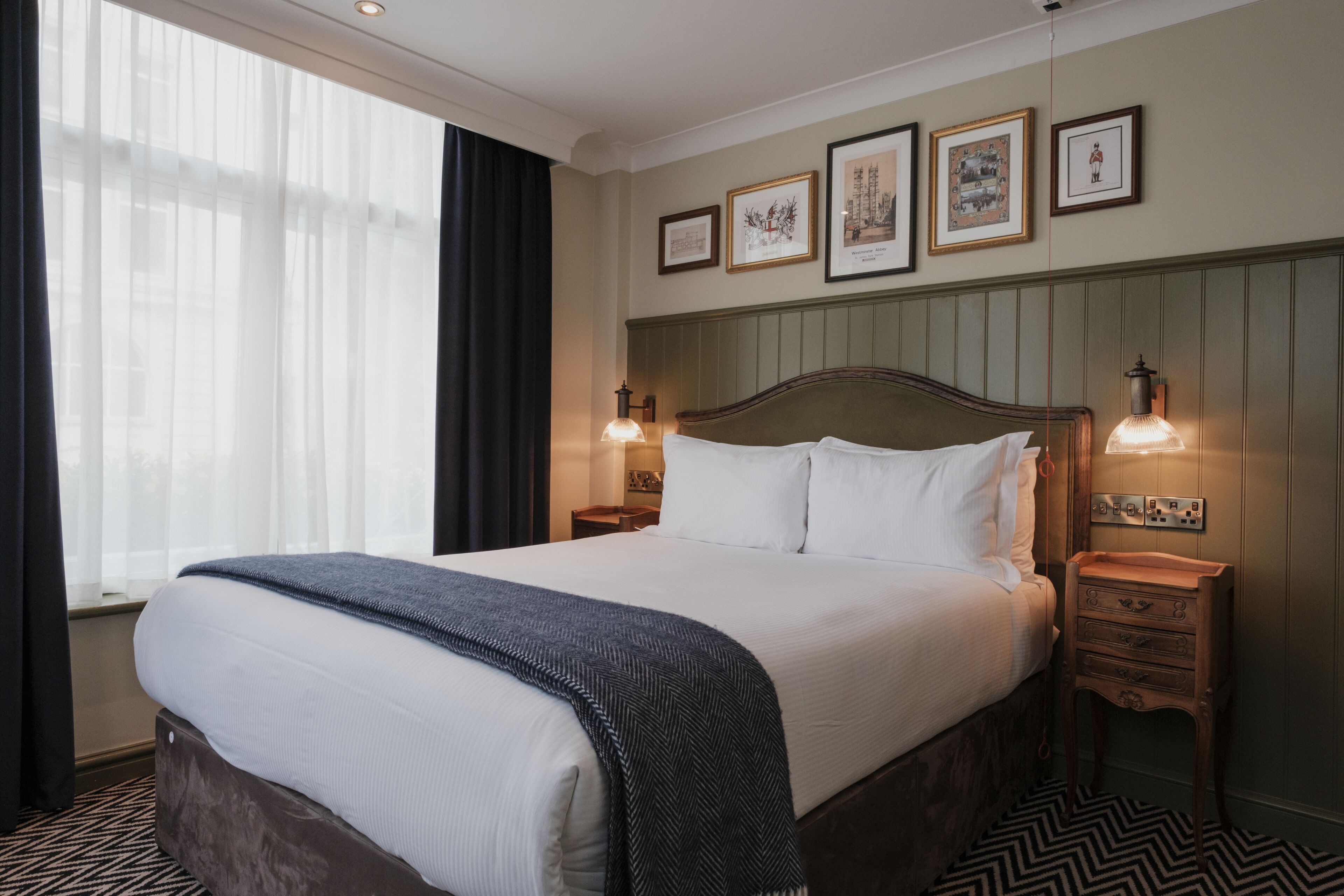 A neatly made bed with a white comforter and a dark throw blanket, a wooden nightstand with a lamp, and a window with sheer curtains.