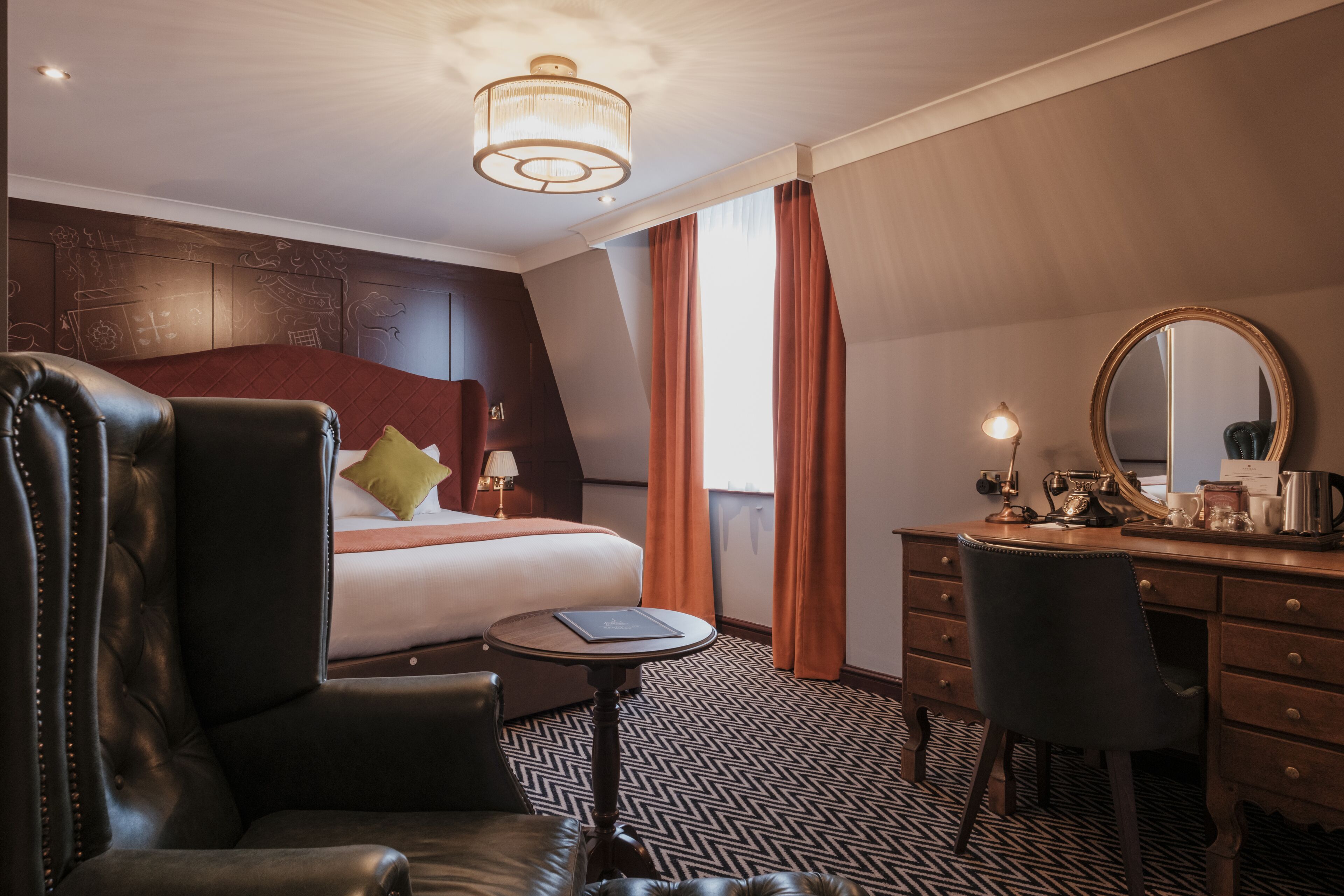 Una camera d'albergo con un letto grande, una scrivania con uno specchio, una poltrona in pelle e un piccolo tavolino rotondo.