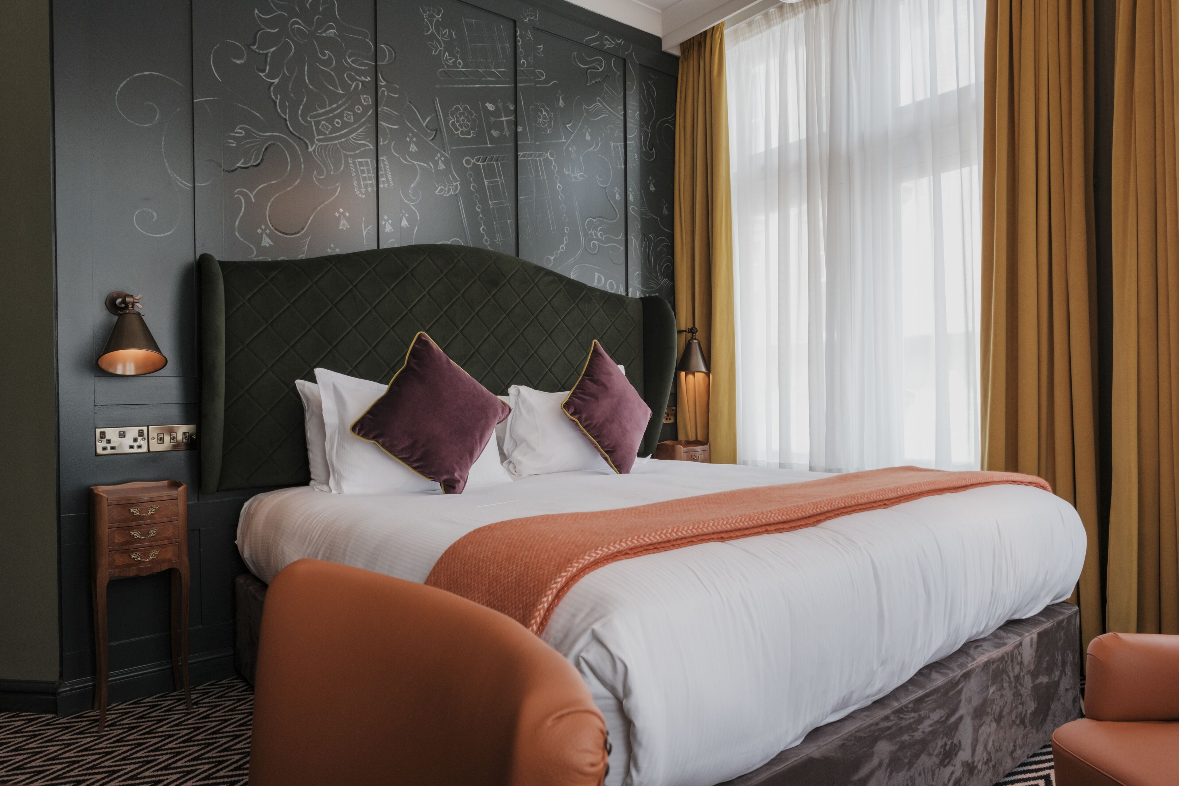 Een hotelkamer met een groot bed, een hoofdeinde met wanddecoratie, een nachtkastje, een lamp en een raam met gordijnen.