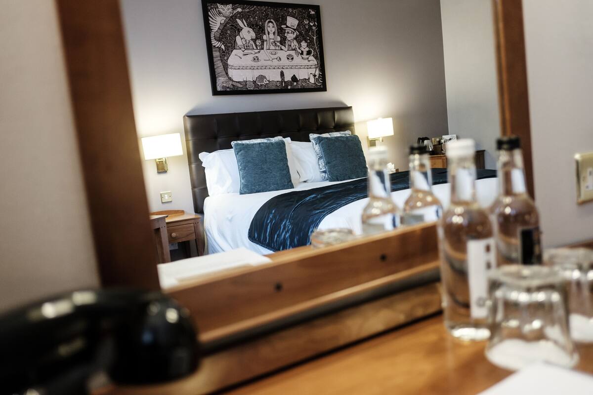 A hotel room with a bed, bedside lamps, a wooden headboard, a framed picture on the wall, and a table with bottles and glasses.