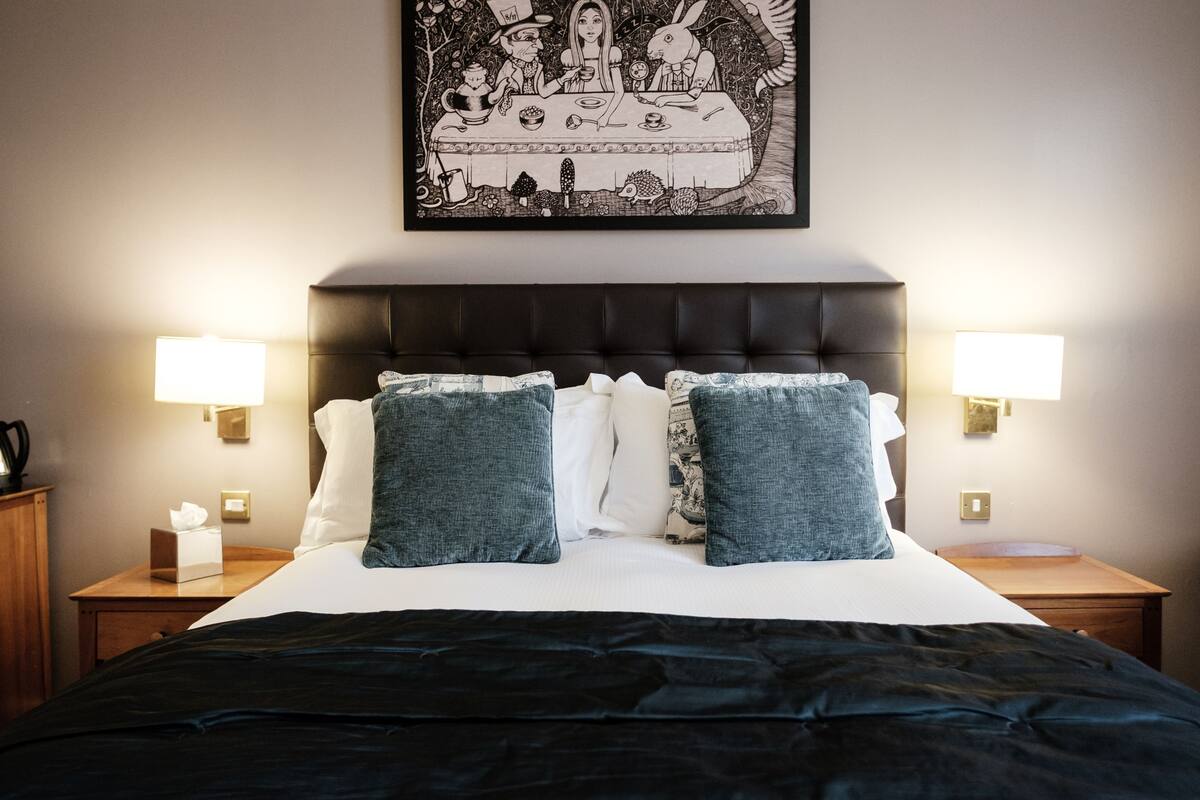 A neatly made bed with a dark cover, two matching pillows, and a framed picture above it. There are two bedside tables with lamps and a small wooden nightstand with a tissue box.