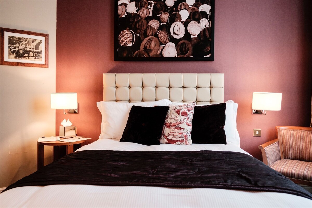 Una habitación de hotel con un cabecero capitonado, una cama grande con ropa de cama blanca y negra, dos lámparas de noche y un cuadro en la pared.